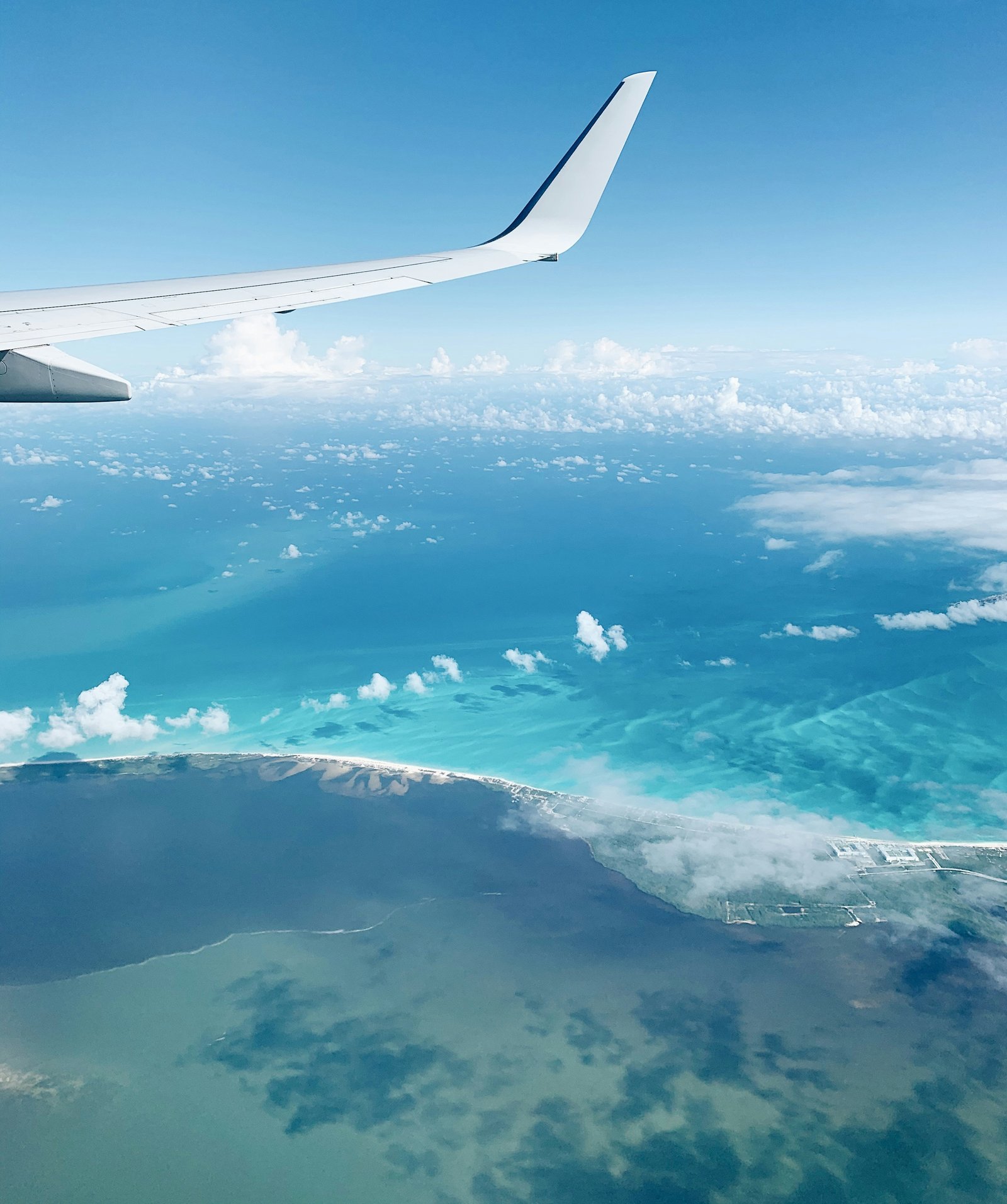 An aircraft wing above a turquoise coastline and scattered clouds.