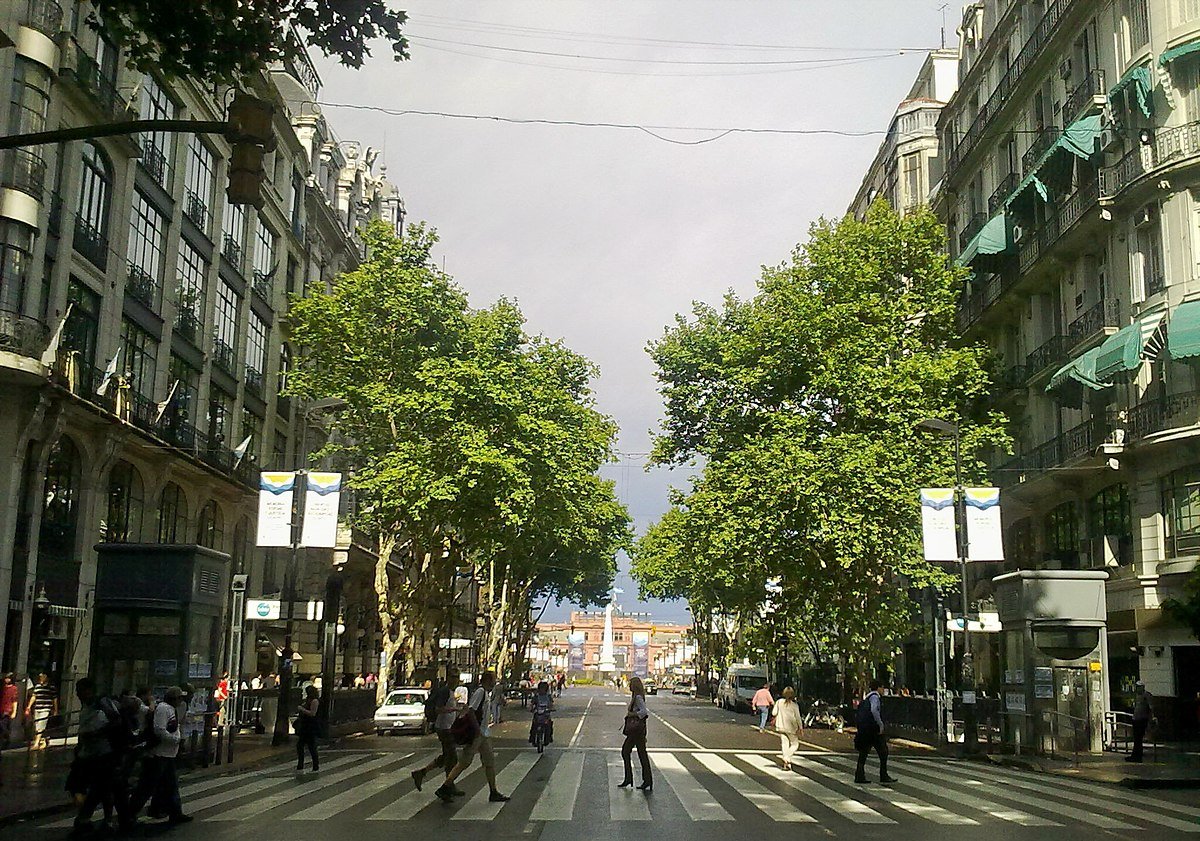 Tree-lined Avenida de Mayo in Buenos Aires with a pedestrian crossing and buildings along the street.
