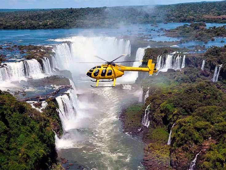 A yellow helicopter flying over Iguazu Falls, with the river and rainforest below.