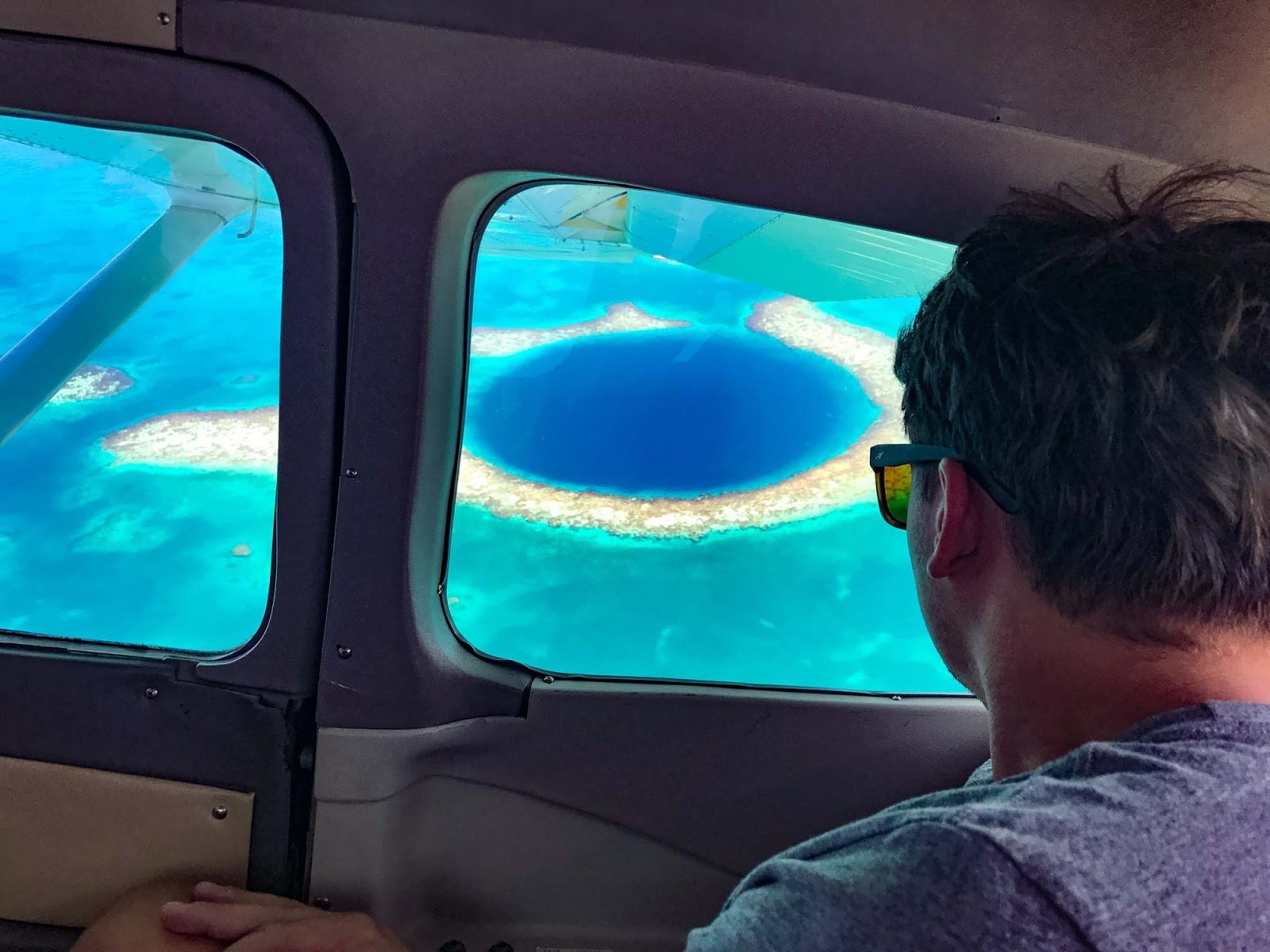View from inside a small aircraft of the Great Blue Hole, a dark blue circle ringed by pale reef and turquoise water.