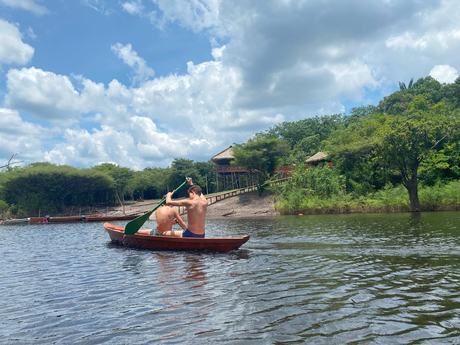 Two paddlers in a canoe on a river beside forest and wooden riverside buildings.