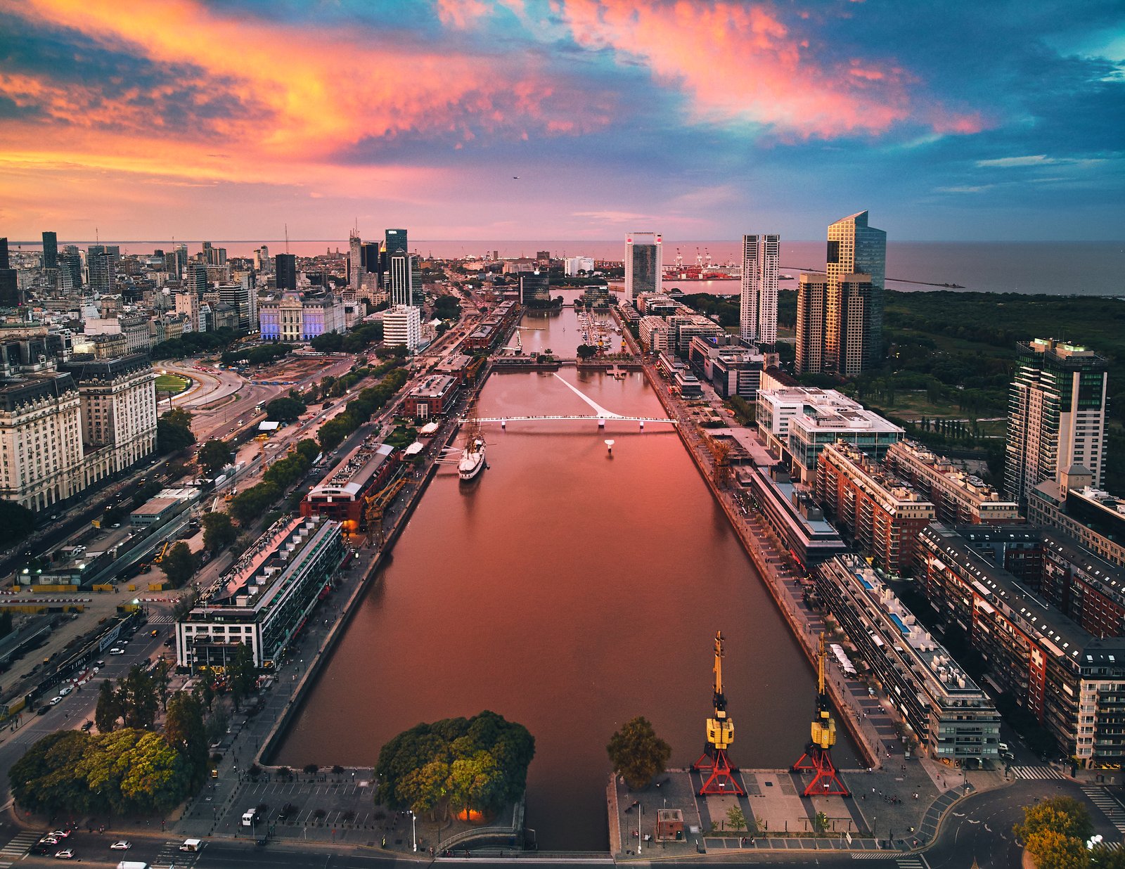Aerial view of Puerto Madero's dock basin and footbridge in Buenos Aires at sunset, with the city's towers along both banks.
