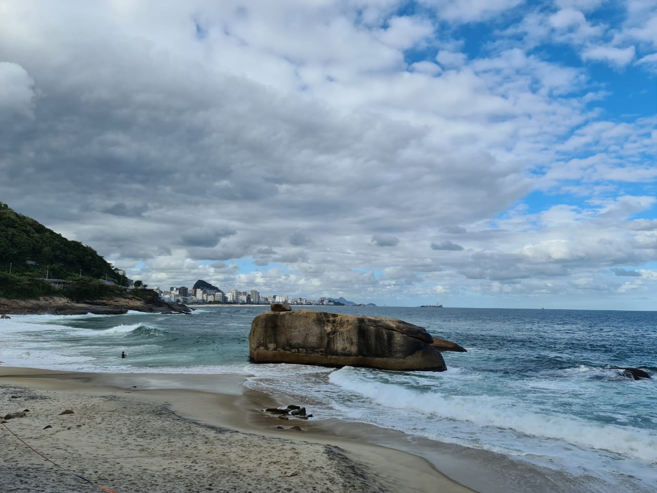Waves breaking around a large rock formation on a Rio de Janeiro beach, with the city skyline visible in the distance