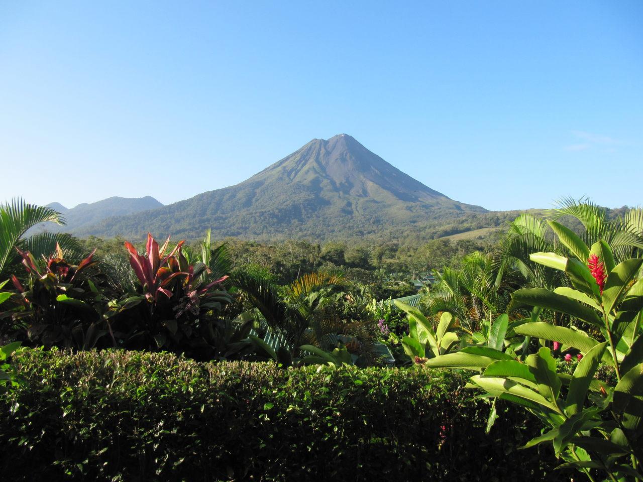 Arenal Volcano rising above green pasture and tropical plants under a blue sky.