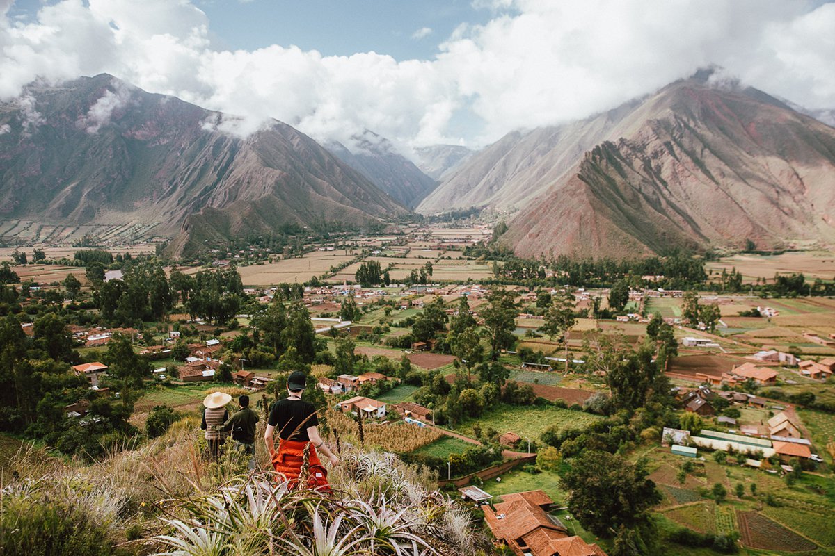 Travellers looking down over Peru's Sacred Valley from a hillside, with terraced fields, a small town and the Andes beyond.