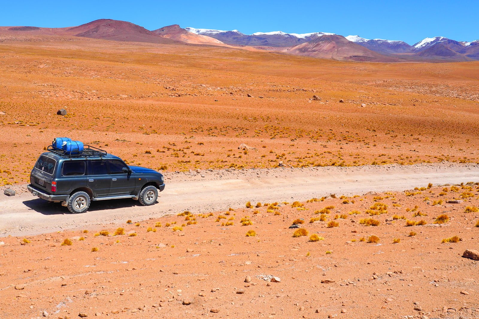 A four-wheel-drive vehicle carrying roof luggage on a road through a red Bolivian landscape