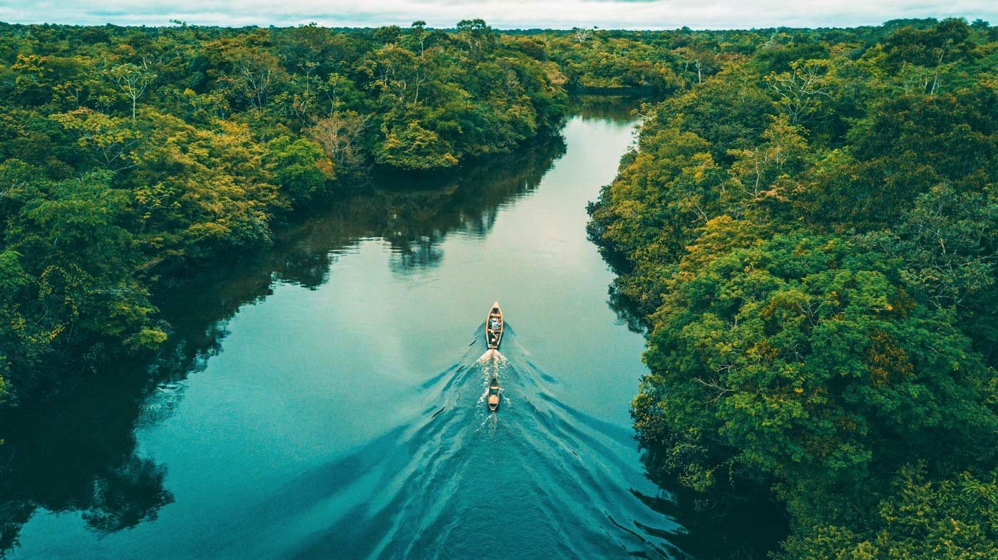 Aerial view of a small boat travelling along a calm Amazon river channel bordered by dense rainforest