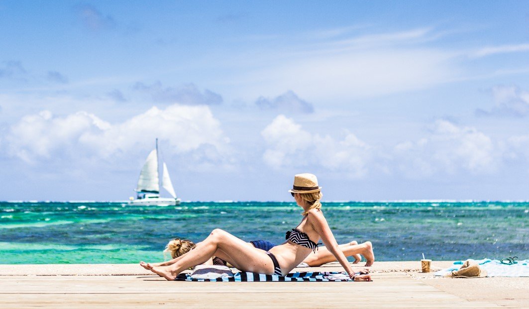 Two visitors resting on a wooden pier beside turquoise water, with a sailboat on the horizon.
