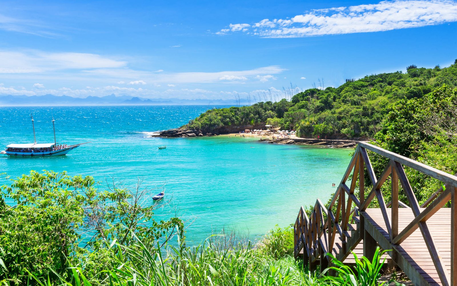 A quiet turquoise cove in Búzios with a wooden viewing platform, a sailing boat and a forested headland