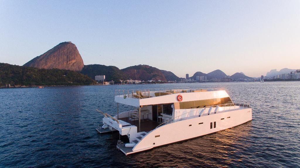 A white private catamaran on Guanabara Bay in Rio de Janeiro with Sugarloaf Mountain behind it at dusk.