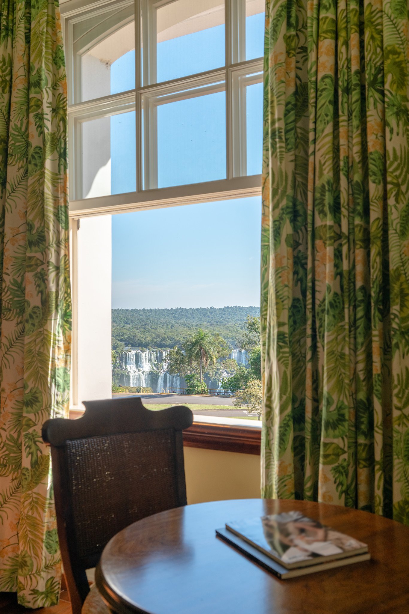 A table and chair beside tall windows framing a view of Iguazu Falls from a hotel room in the forest.