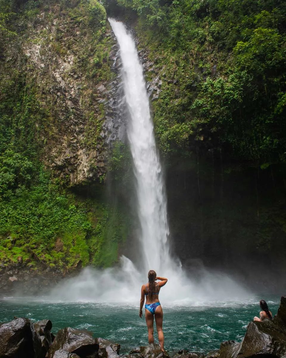 A waterfall dropping into a green pool in Costa Rican forest, with one visitor standing at the water's edge.